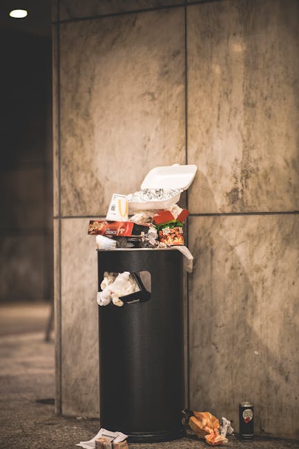 A black cylindrical public waste bin situated against a large beige tiled wall in an indoor setting. The top of the bin is open and overfilled with assorted fast food packaging, including paper carton