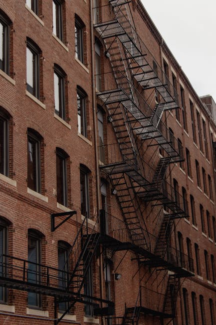 A close-up view of an exterior brick building showing multiple black metal fire escapes attached to the façade. The building features rows of evenly spaced rectangular windows with black frames, some with open or closed shutters. The daylight illuminates the brick surface, highlighting its reddish-brown tones and textured surface. The fire escapes consist of staircases and platforms with railings, zigzagging down the side of the building. The scene depicts an urban residential setting, with no cleaning tools or surfaces visible in this exterior shot. This image is relevant to surface cleaning and maintenance in residential buildings, as relevant to Pimlico lofts and mould remediation, with a focus on building hygiene upkeep. For professional cleaning or sanitisation, please contact Cleaners Pimlico as featured on the page.