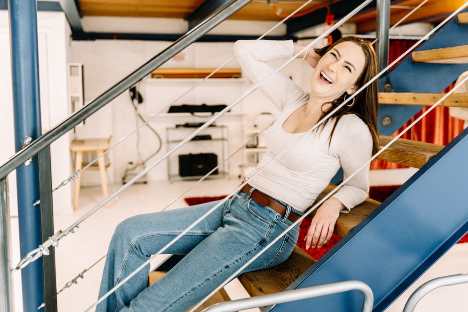 A young woman with long brown hair, wearing a light grey long-sleeve shirt and blue jeans, is seated on a wooden staircase with metal railings and rope balustrades. She is smiling broadly and appears to be enjoying herself, with her right arm behind her head and her left hand resting on the stairs. The staircase is located in a modern, well-lit room with white walls, a tiled floor, and visible wooden accents. A red and blue slide is attached to the staircase, suggesting a playful, residential environment. The background includes a small wooden bench, a black radiator, and some shelving, all presented in a clean and tidy condition. The lighting is bright and natural, enhancing the scene’s cheerful atmosphere. The image represents a domestic setting with a focus on cheerful leisure and well-maintained interiors, consistent with the themes of cleaning and home hygiene promoted by Cleaners Pimlico.