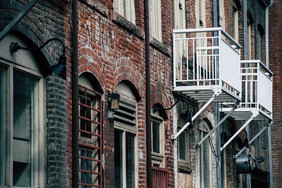 A close-up view of an exterior brick building showing multiple black metal fire escapes attached to the façade. The building features rows of evenly spaced rectangular windows with black frames, some with open or closed shutters. The daylight illuminates the brick surface, highlighting its reddish-brown tones and textured surface. The fire escapes consist of staircases and platforms with railings, zigzagging down the side of the building. The scene depicts an urban residential setting, with no cleaning tools or surfaces visible in this exterior shot. This image is relevant to surface cleaning and maintenance in residential buildings, as relevant to Pimlico lofts and mould remediation, with a focus on building hygiene upkeep. For professional cleaning or sanitisation, please contact Cleaners Pimlico as featured on the page.