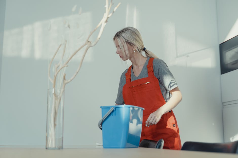 Two professional cleaners from Cleaners Pimlico standing indoors against a plain white wall, dressed in beige uniforms with short sleeves and white collars. The cleaner on the left, an Asian woman wit