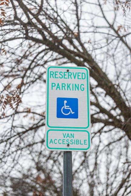 A reserved parking sign for disabled and van accessibility mounted on a metal pole, set against a background of leafless tree branches and an overcast sky. The sign features a blue and white wheelchair icon, with the text 'RESERVED PARKING' at the top and 'VAN ACCESSIBLE' at the bottom, outlined with a green border. The image emphasizes outdoor signage related to accessible parking policies, relevant to cleaning and maintenance of parking area signage, as part of compliance with local regulations. For professional cleaning of such signage or surrounding surfaces, [COMPANY_NAME] at cleaners-pimlico.com offers specialist services to ensure clarity and visibility.