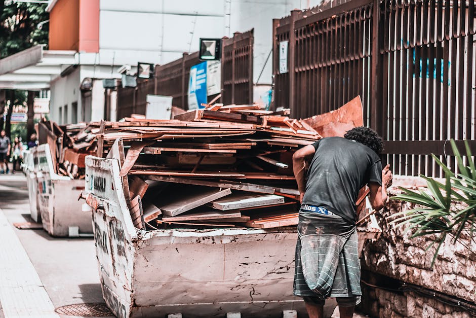 A man with curly hair, wearing a dark t-shirt and checkered shorts, is seen from behind as he examines a large, overflowing dumpster filled with various construction and renovation waste, including wooden planks, tiles, and debris, situated on a city street adjacent to a brown metal fence. In the background, there are buildings, signage, and blurred pedestrians, with the scene illuminated by natural daylight. The image highlights waste disposal related to renovation projects, relevant to regulations about skip permits and waste management in Pimlico, as covered by Cleaners Pimlico on their cleaning service page.
