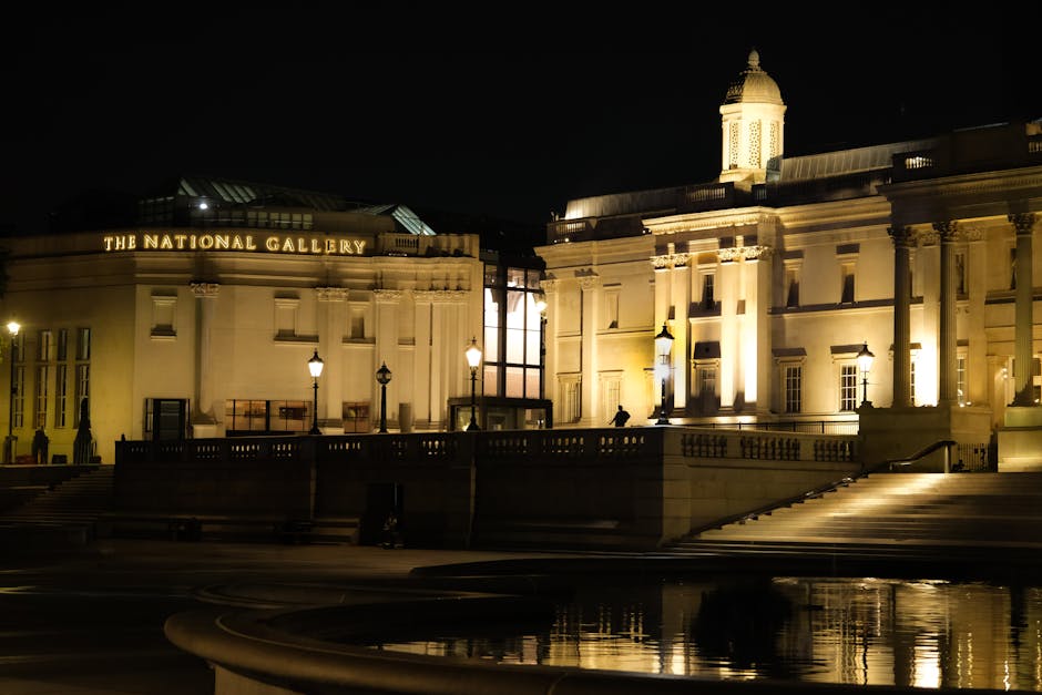 Night view of the illuminated exterior of the National Gallery building in Tate Britain, showing its classical architecture with large columns and ornate details, softly lit by exterior lighting. The stone surfaces are clean and polished, reflecting the warm light, with a smooth stone staircase leading up to the entrance. The surrounding area is quiet and tidy, with a few lamp posts casting light on the paved ground. The lighting emphasizes the building's historical features, and the scene appears well-maintained, indicative of professional cleaning and upkeep by Cleaners Pimlico.