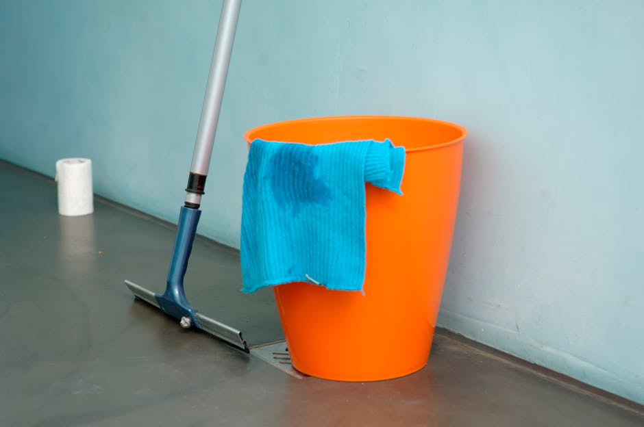 An image of a bright orange plastic bucket placed on a smooth, gray floor next to a wall, with a blue microfiber cleaning cloth draped over the side of the bucket showing dampness. A mop with a blue handle and a flat cleaning head is leaning against the wall nearby, positioned over a drain on the floor. In the background, a partially visible roll of white toilet paper or paper towels is on the floor. The room appears to be well-lit and clean, suggesting an area undergoing surface cleaning or spill clean-up. The scene is indicative of professional sanitisation or deep cleaning activities, as performed by Cleaners Pimlico, servicing Pimlico venues and the Tate Britain area.