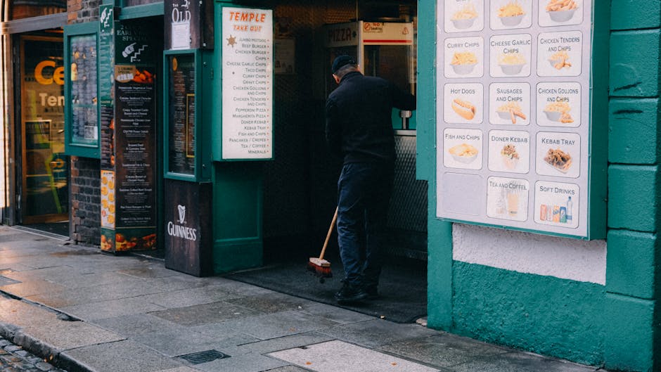 A commercial cleaning worker dressed in dark clothing, including a black cap and jacket, is using a push broom to sweep the entrance area of a restaurant on Warwick Way. The restaurant's exterior features bright signage displaying various takeaway food options such as chips, kebabs, and hot dogs, on a large white menu board mounted on a teal-painted wall. The ground is wet and appears to have been recently cleaned, with water reflecting the surrounding shopfronts. To the left, another establishment's window and door are visible, indicating a busy street environment. The scene is well-lit by natural daylight, emphasizing the cleanliness and organized appearance of the entrance space, which is maintained by the professional team at Cleaners Pimlico focusing on surface cleaning and hygiene for commercial properties.