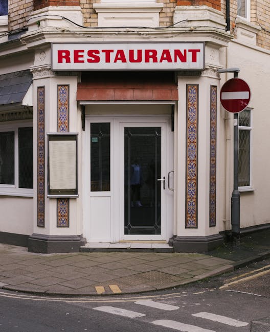 A commercial cleaning worker dressed in dark clothing, including a black cap and jacket, is using a push broom to sweep the entrance area of a restaurant on Warwick Way. The restaurant's exterior features bright signage displaying various takeaway food options such as chips, kebabs, and hot dogs, on a large white menu board mounted on a teal-painted wall. The ground is wet and appears to have been recently cleaned, with water reflecting the surrounding shopfronts. To the left, another establishment's window and door are visible, indicating a busy street environment. The scene is well-lit by natural daylight, emphasizing the cleanliness and organized appearance of the entrance space, which is maintained by the professional team at Cleaners Pimlico focusing on surface cleaning and hygiene for commercial properties.